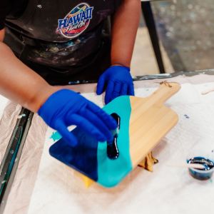 Person painting a wooden board with blue paint while wearing gloves.