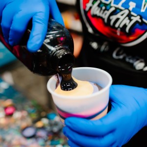 Person pouring black paint into a layered cup, wearing blue gloves.