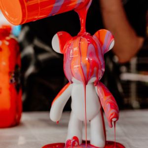 Person pouring orange and purple paint over white bear figurine.