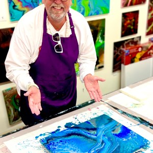 Smiling man in purple apron shows his abstract blue painting in a colorful art studio.